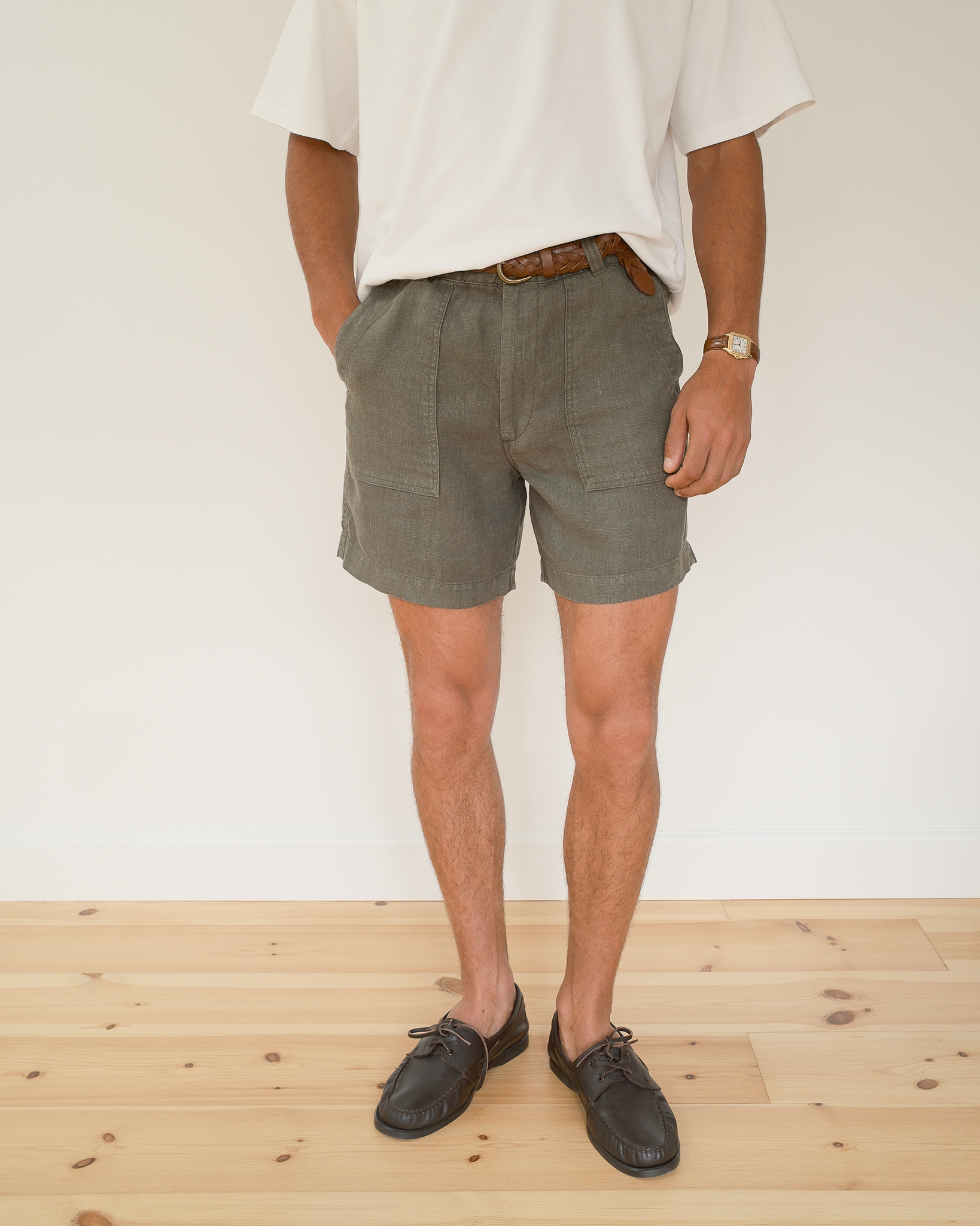A person wearing olive green linen shorts, a white t-shirt, a brown leather belt, and brown leather shoes, standing against a white wall on a light wood floor.