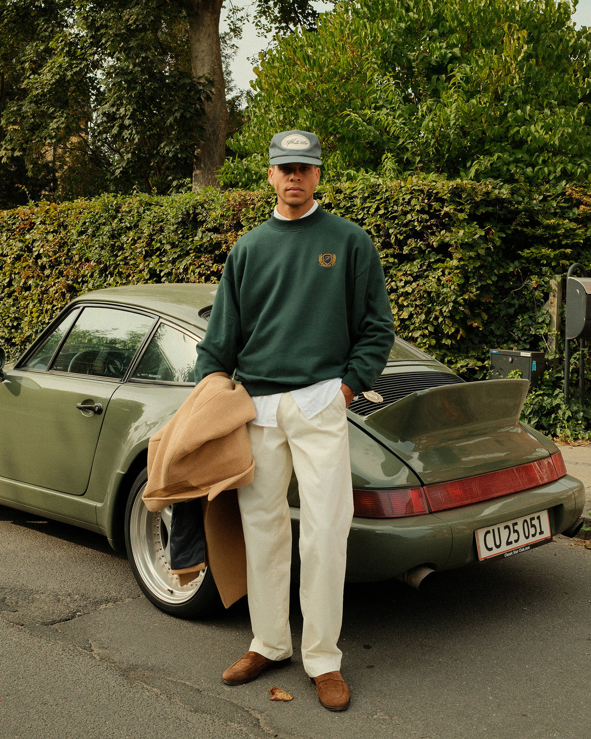 A man stands outdoors in front of a vintage olive green Porsche, wearing a dark green crewneck sweatshirt, cream trousers, brown loafers, and holding a tan jacket. The sweatshirt features a small embroidered logo on the left chest.