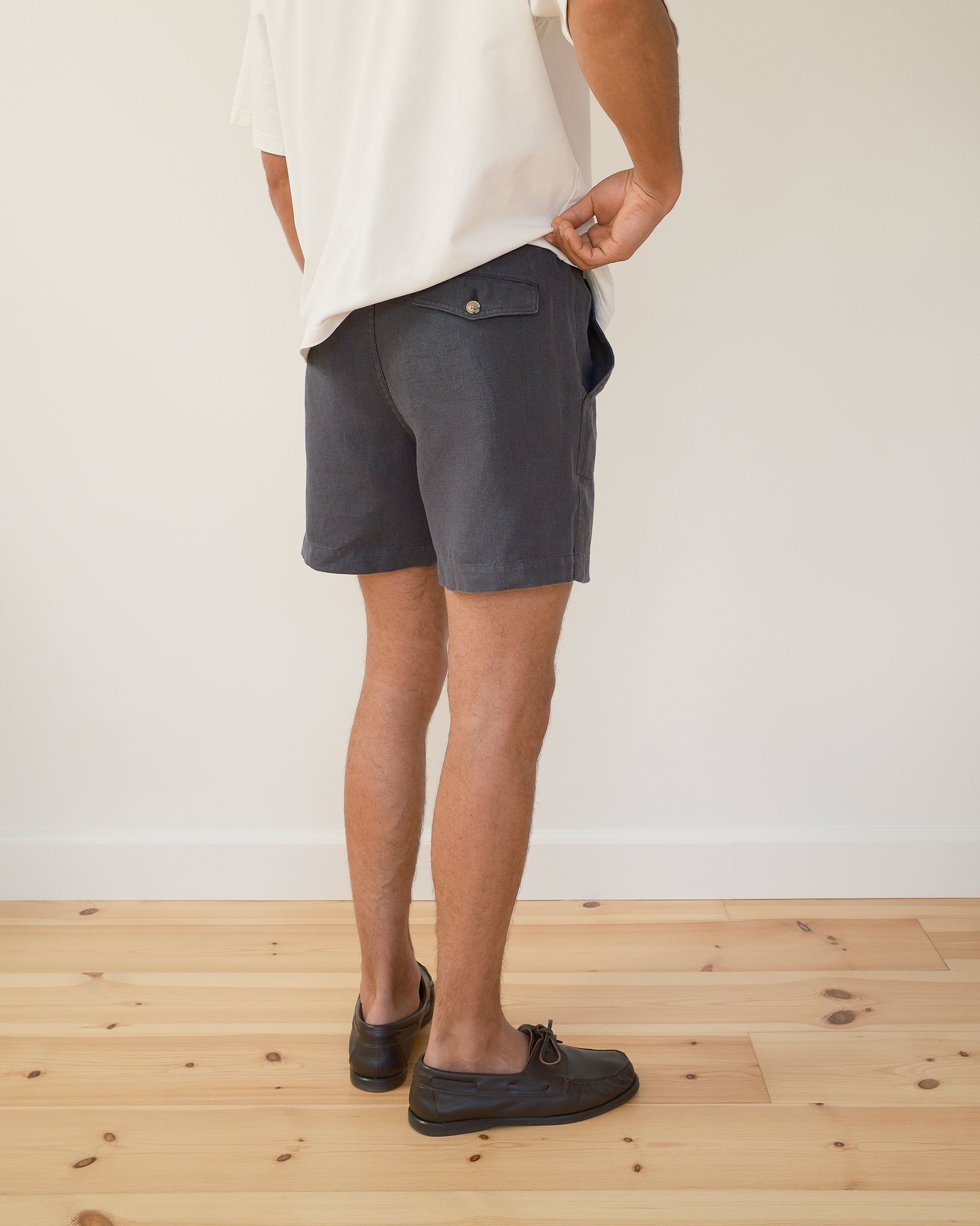 A person shown from the waist down wearing dark blue shorts, a white t-shirt, and boat shoes, standing on a light wood floor against a white wall.