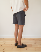 A person shown from the waist down wearing dark blue shorts, a white t-shirt, and boat shoes, standing on a light wood floor against a white wall.