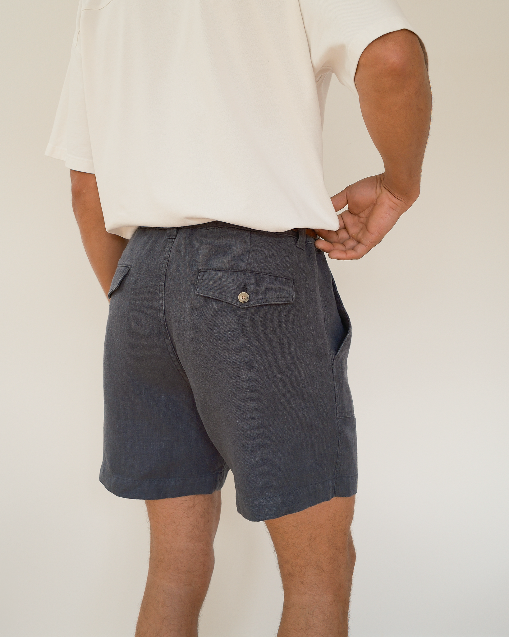 A person shown from the waist down wearing dark blue shorts, a white t-shirt,  standing on a light wood floor against a white wall.