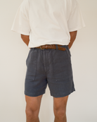 A person shown from the back wearing dark blue linen shorts with a brown woven belt, paired with a white t-shirt, against a plain off-white background.