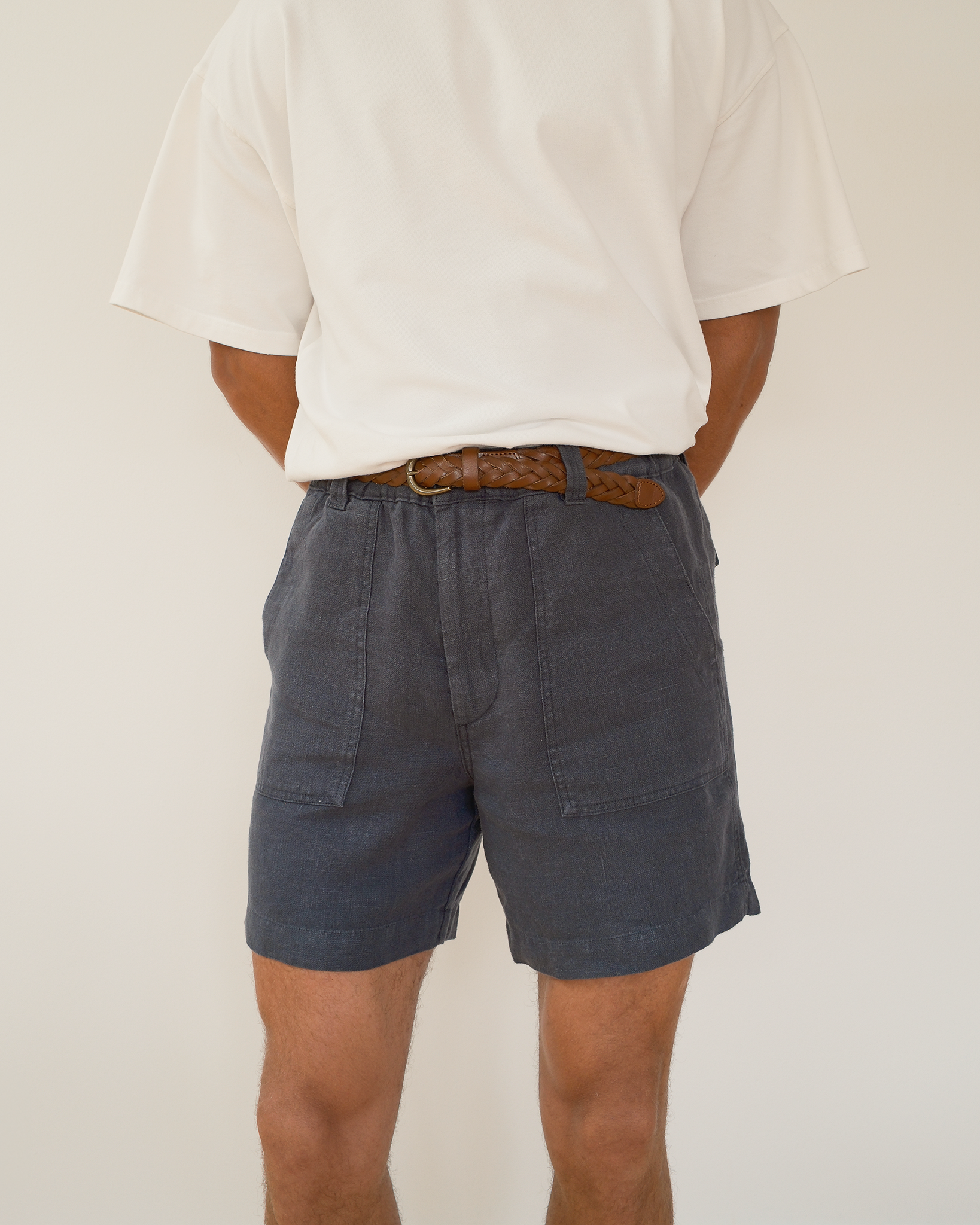 A person shown from the back wearing dark blue linen shorts with a brown woven belt, paired with a white t-shirt, against a plain off-white background.