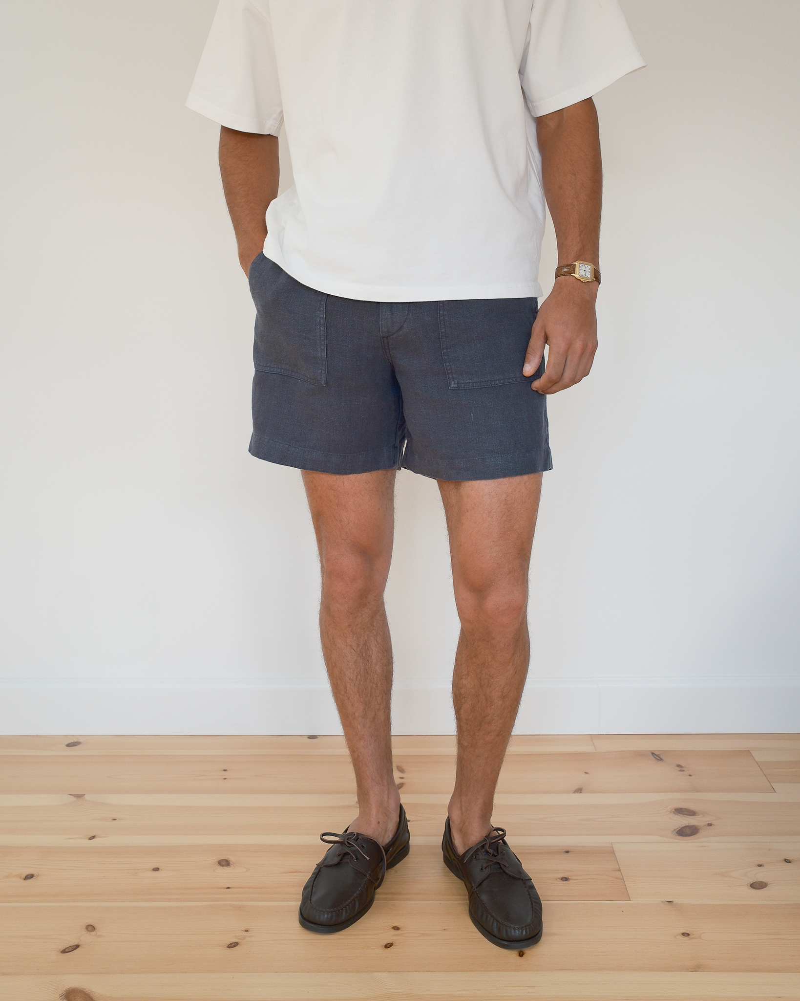 A person standing indoors wearing dark blue linen shorts, a white t-shirt, and dark brown leather shoes on a light wood floor against a white wall.