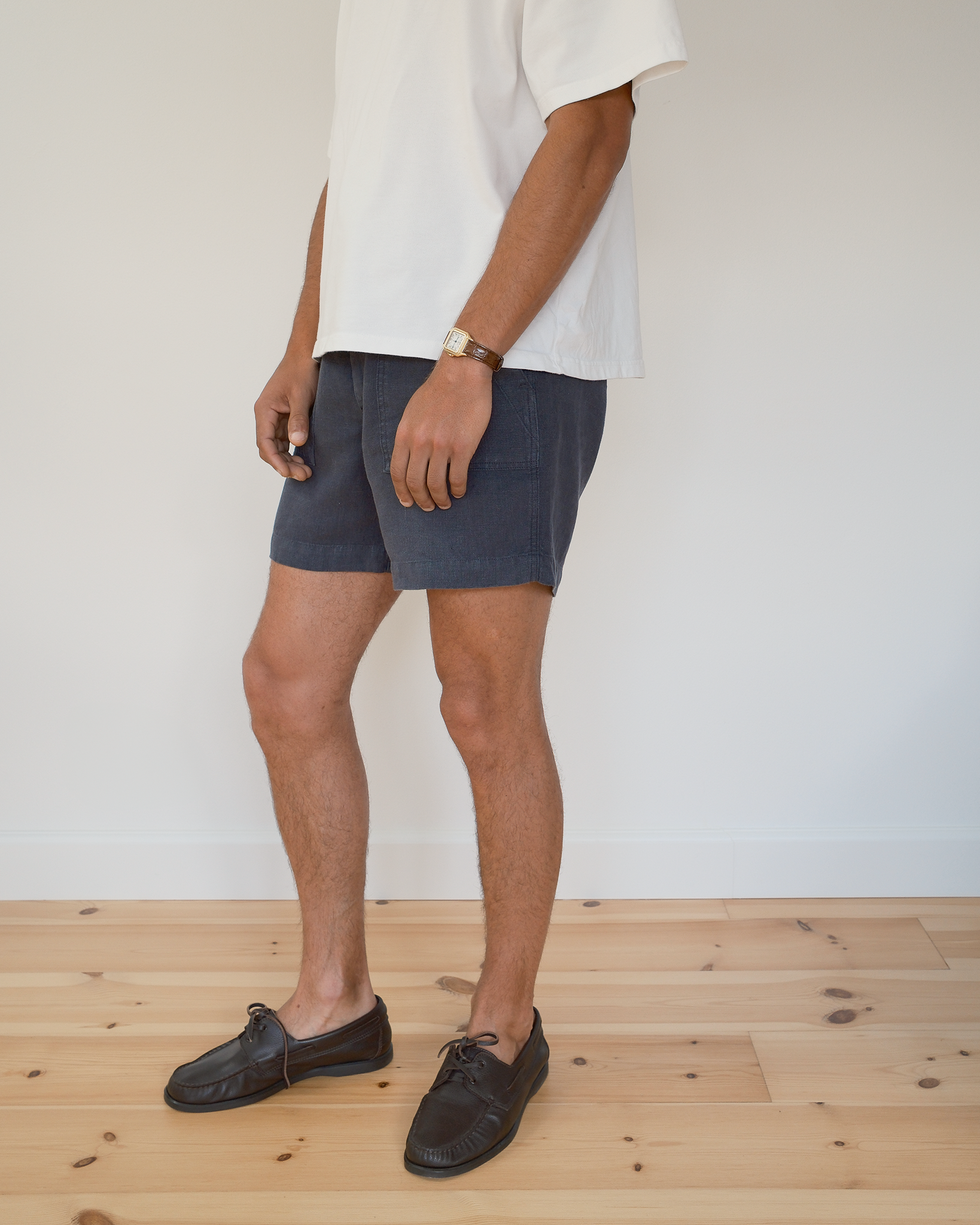 A person wearing dark blue shorts, a white t-shirt, and dark brown boat shoes, standing on a light wood floor against a white wall.