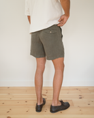 A person shown from the back wearing olive green shorts, a white t-shirt, and black shoes, standing on a light wood floor against a white wall.