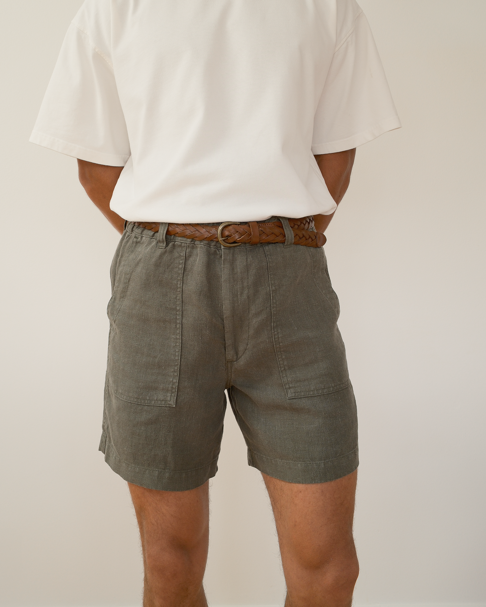 A person shown from the back wearing olive green linen shorts, a white t-shirt, and a brown woven belt against a plain off-white background.