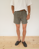 A person wearing olive green linen shorts, a white t-shirt, a brown leather belt, and brown leather shoes, standing against a white wall on a light wood floor.
