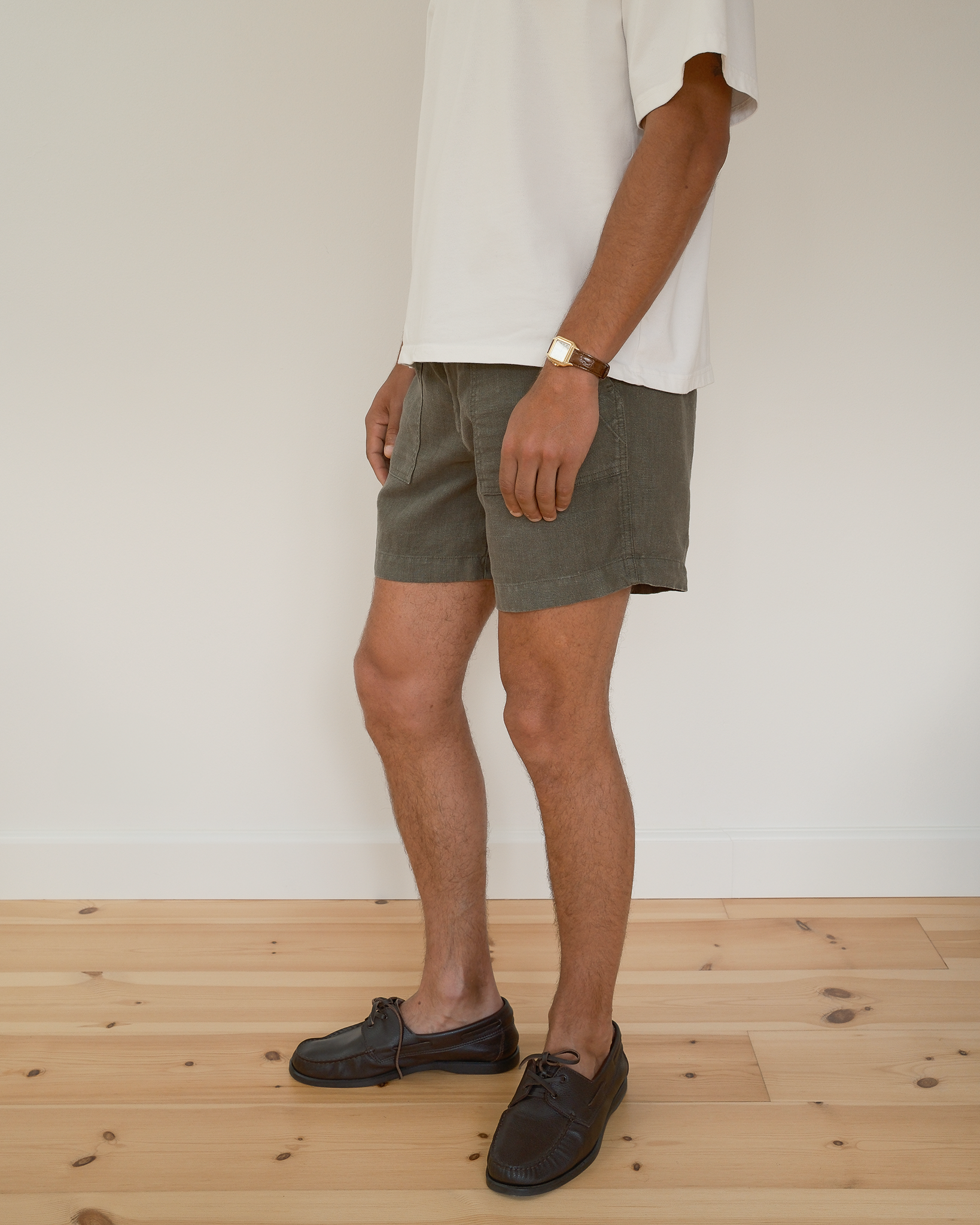 A person wearing olive green shorts, a white t-shirt, and dark brown boat shoes, standing on a light wood floor against a white wall.