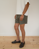 A person wearing olive green shorts, a white t-shirt, and dark brown boat shoes, standing on a light wood floor against a white wall.