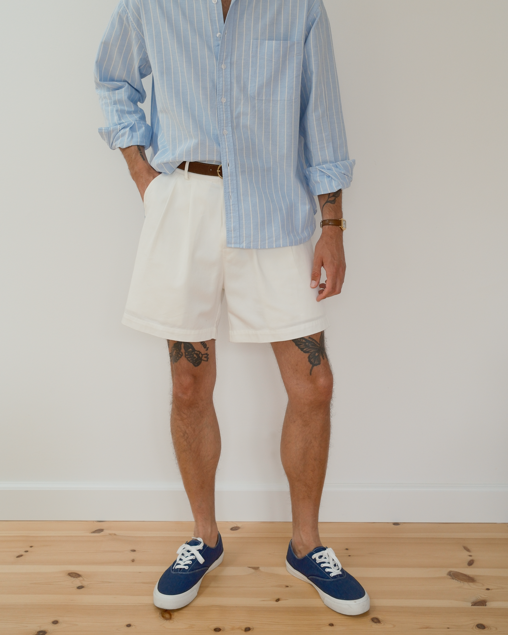 A man wearing a light blue and white vertically striped button-down shirt, off-white shorts, a brown belt, and blue canvas sneakers, standing against a white wall on a light wood floor.