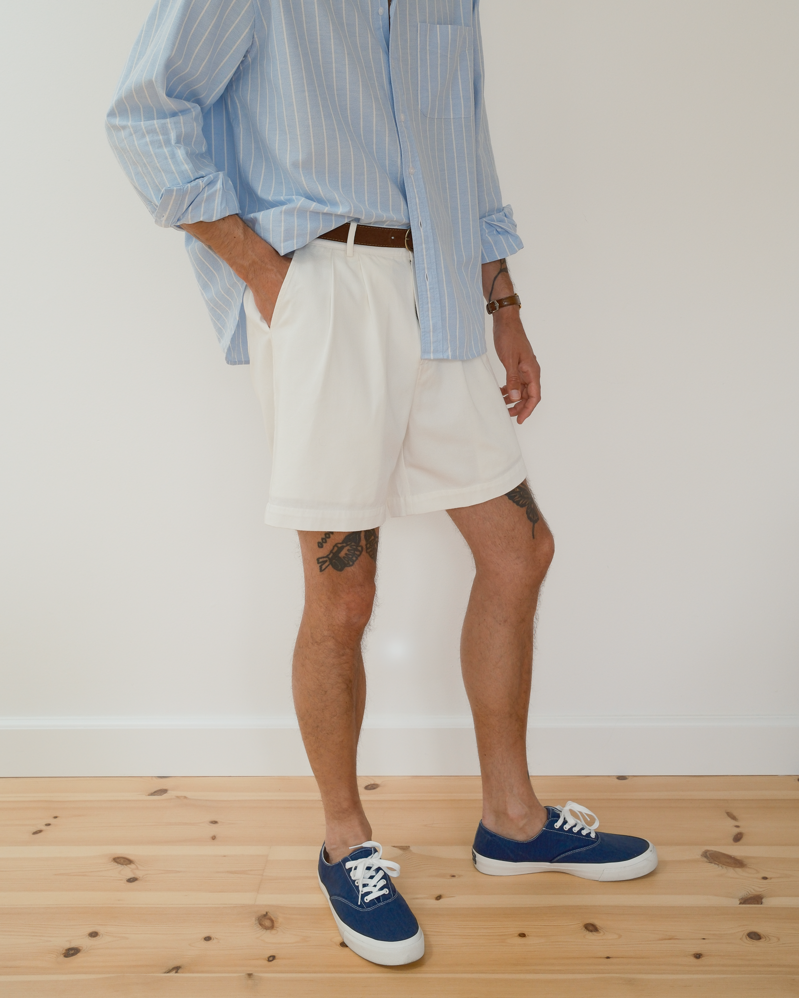 A person wearing white pleated shorts, a light blue and white striped button-down shirt, and blue canvas sneakers, standing on a light wood floor against a white wall.