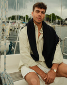 man wearing a pistachio green button down oxford shirt white shirts and a sweater over the shoulders while sitting on a boat. 
