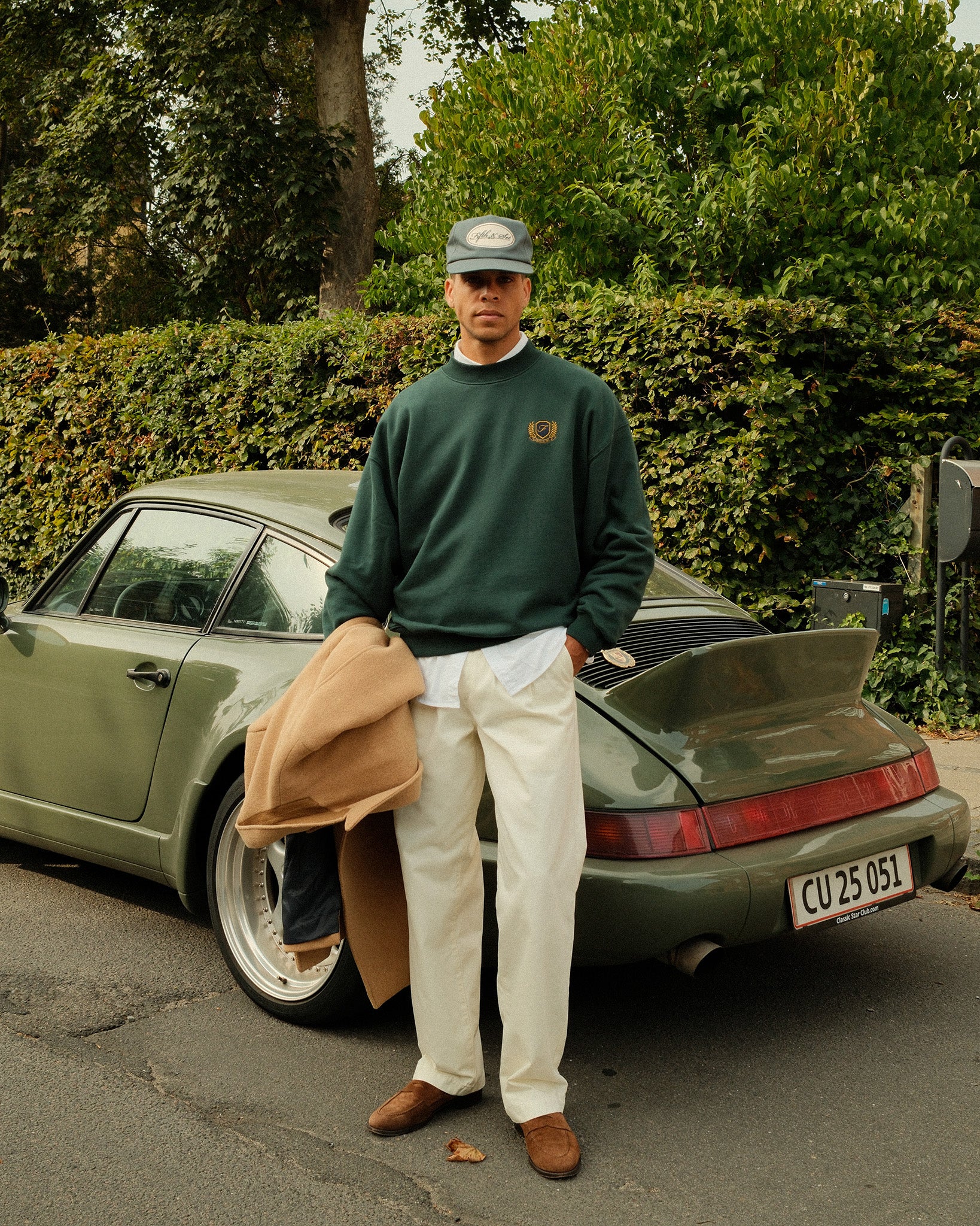 A man stands outdoors in front of a vintage olive green Porsche, wearing a dark green crewneck sweatshirt, cream trousers, brown loafers, and holding a tan jacket. The sweatshirt features a small embroidered logo on the left chest.