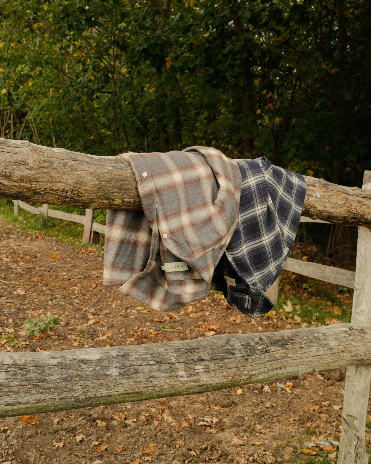 Two plaid flannel shirts, one in beige and brown and one in navy and white, hanging from a rustic wooden fence in an outdoor wooded setting.