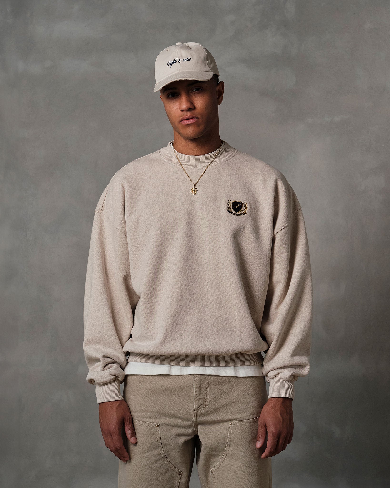 Male model wearing oatmeal colored sweatshirt, khaki pants and beige cap.